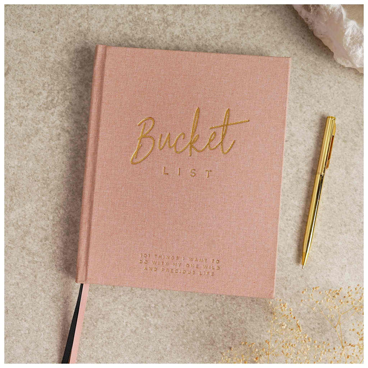 Pink notebook titled 'Bucket List' with a gold pen on a textured surface